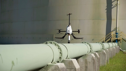 A quadcopter drone hovers above a large industrial pipeline supported by concrete stands near a storage tank