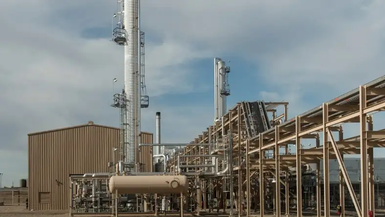 A tan industrial building and horizontal storage tank sit beside tall towers and elevated pipe racks