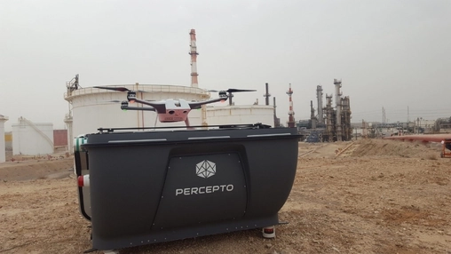 Quadcopter drone hovering above a matte black docking station at an outdoor industrial refinery site