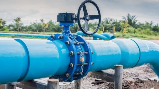 A padlocked valve on a large blue pipeline with a metal control wheel and greenery in the background