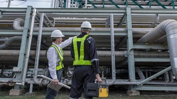 Two workers in safety gear inspect industrial piping while carrying tools at a processing plant