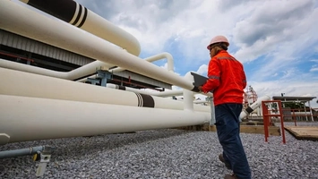 Worker in safety gear inspects industrial pipelines while carrying a laptop at a facility