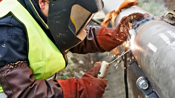A worker wearing thick leather gloves welds a large metal pipeline as bright orange sparks scatter outward
