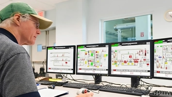 Man in a green cap seated at a control room desk viewing four monitors displaying pipeline schematics
