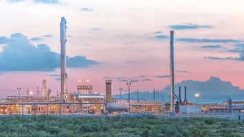 Industrial gas processing facility with tall flare stacks silhouetted against a pink evening sky above