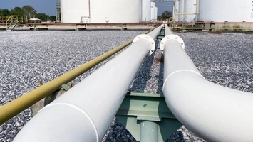 Two large white industrial pipes converge across a gravel yard leading toward storage tanks in the distance