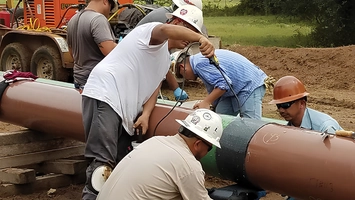 Crew of workers in safety gear working on a section of large diameter pipe near trees and heavy equipment