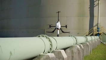 A quadcopter drone hovers above a large industrial pipeline supported by concrete stands near a storage tank