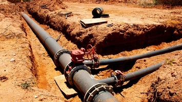 Steel pipeline fittings and red valve wheels visible in an open dirt trench during installation work