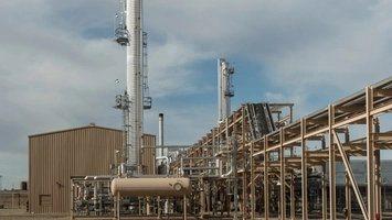 A tan industrial building and horizontal storage tank sit beside tall towers and elevated pipe racks