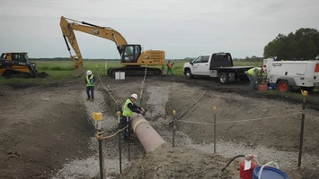 Workers in hard hats lower a large pipe into a gravel-lined trench beside an excavator and pickup truck