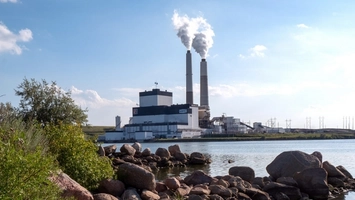 Coal power station with steam from paired smokestacks, viewed past rocks and greenery at the water's edge